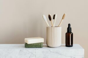 Collection of organic soaps and bamboo toothbrushes in ceramic minimalism style holder placed near renewable glass bottle with essential oil on white marble tabletop against beige wall